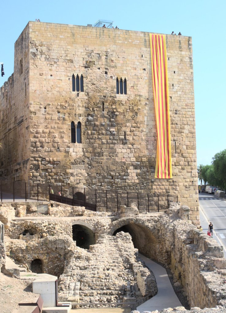 Старинная башня, с неё свисает длинный полосатый каталонский флаг