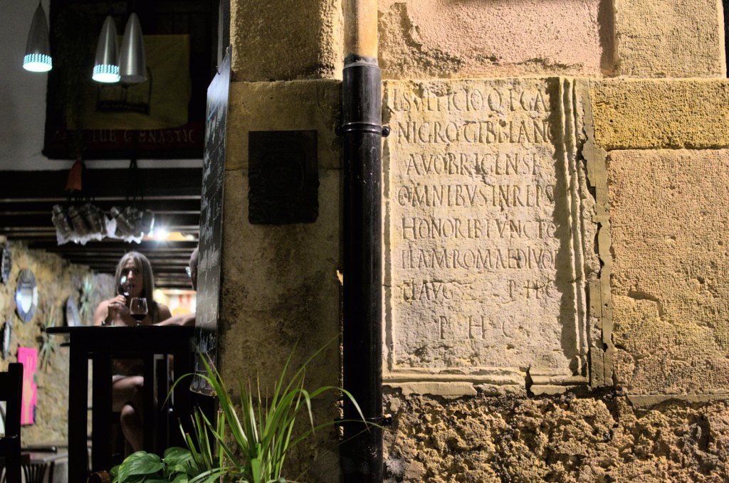 В дверной проем видны посетители бара; справа от двери — плита с латинской надписью.