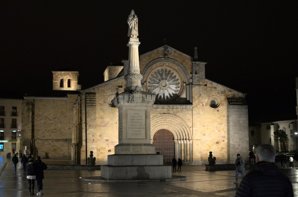 Вечерний вид площади; на дальнем плане — церковь с романским арочным порталом, ближе — памятник в виде человеческой фигуры на колонне