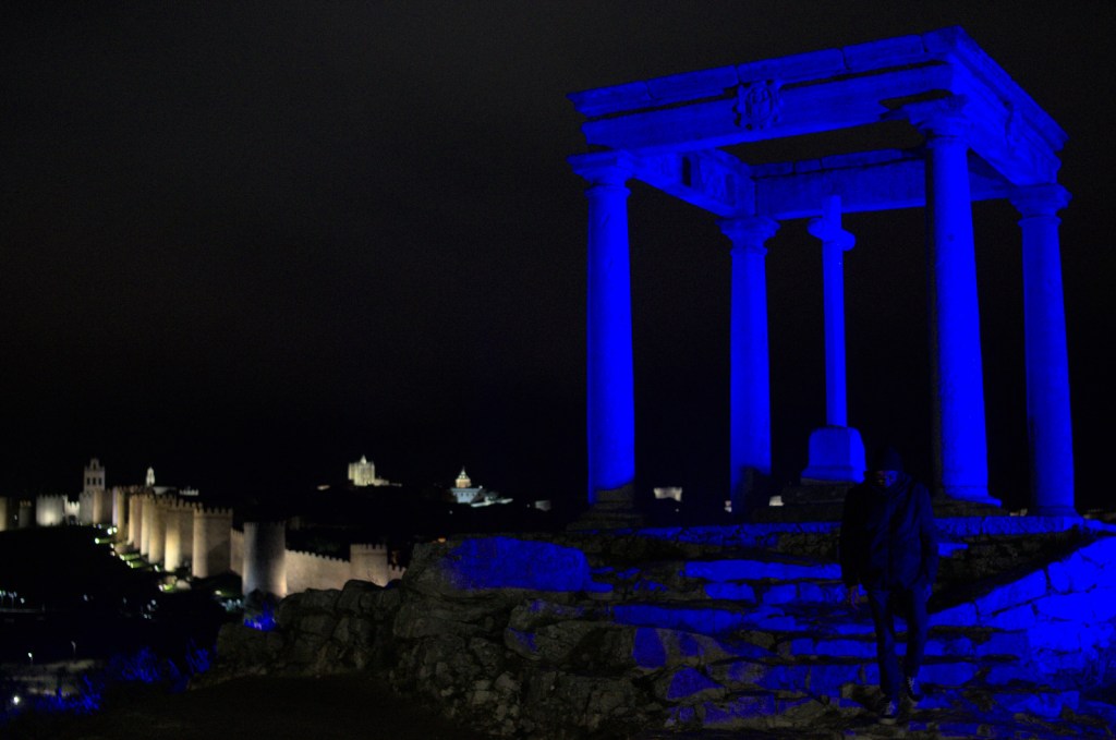 Постройка в виде карниза, опирающегося на четыре колонны, подсвечена синим светом; вдалеке — фрагмент городских стен с тёплым тоном подсветки