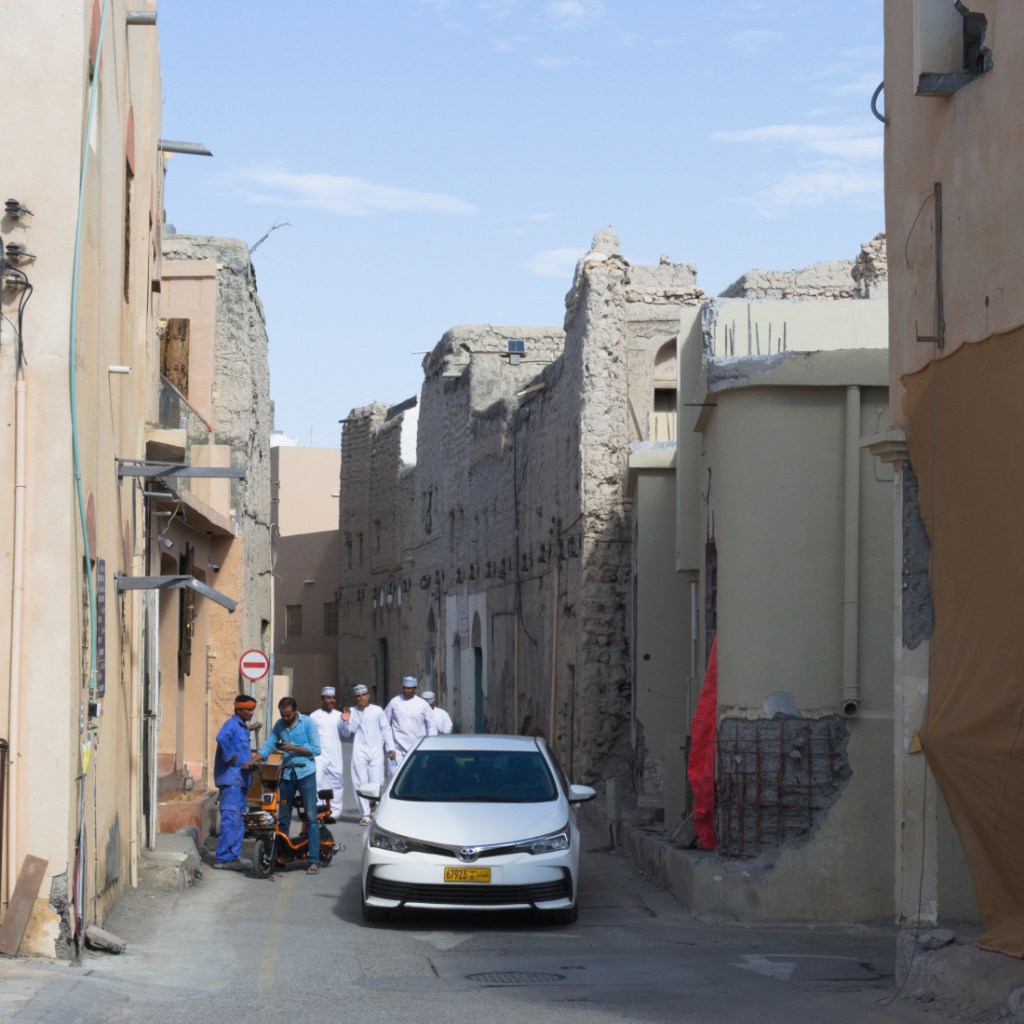 Рабочие, прохожие и автомобиль на узкой улице между глинобитных домов