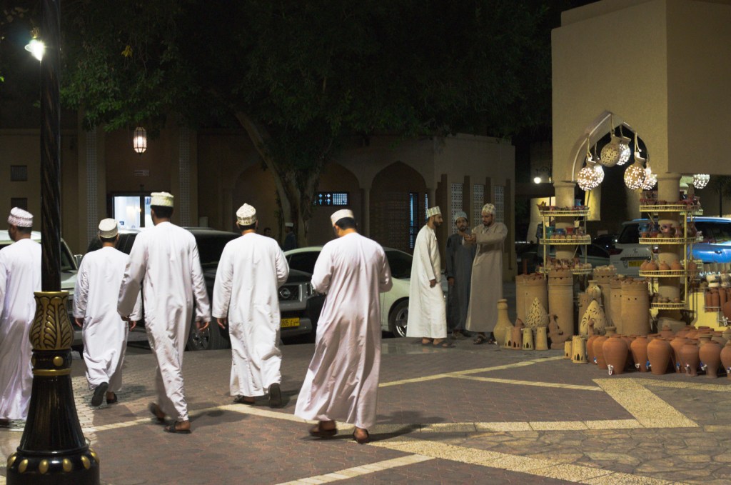 Мужчины в белых оманских нарядах идут по рынку в вечерней темноте
