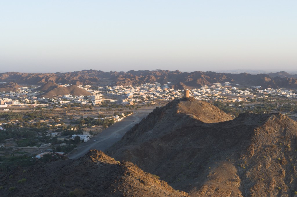 Складки гор в лучах вечернего солнца. Белые дома у склонов гор. Сторожевая башня на вершине.