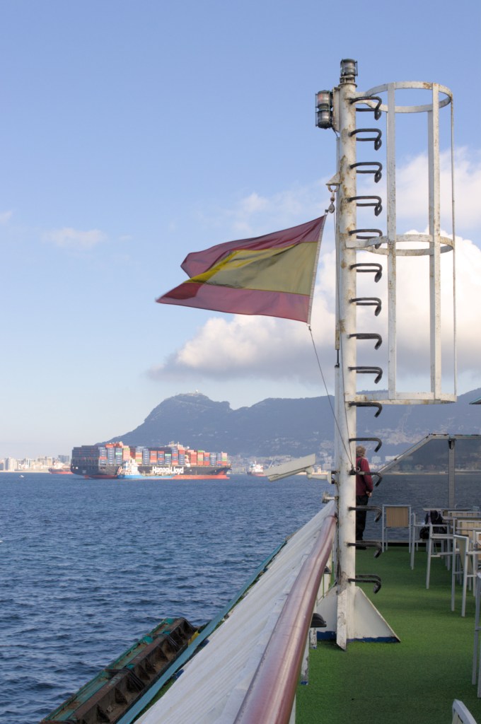 Испанский флаг на судне, Гибралтарская скала и контейнеровоз вдалеке.