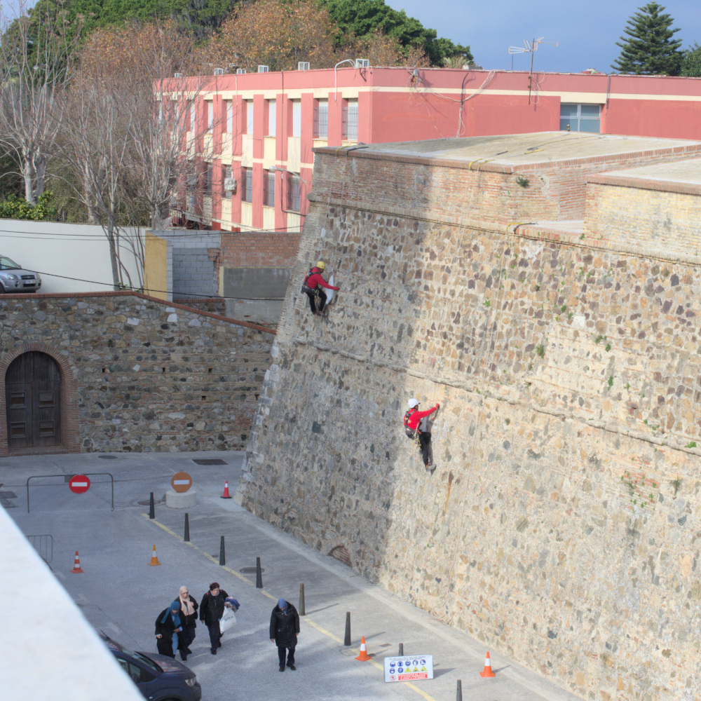 Рабочие выполняют высотные работы на стене, внизу по улице идёт группа женщин