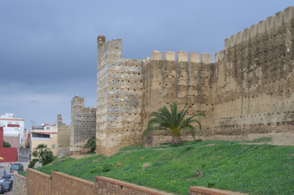 Остатки стен, вдоль них зелёная трава, вдали небольшие жилые дома